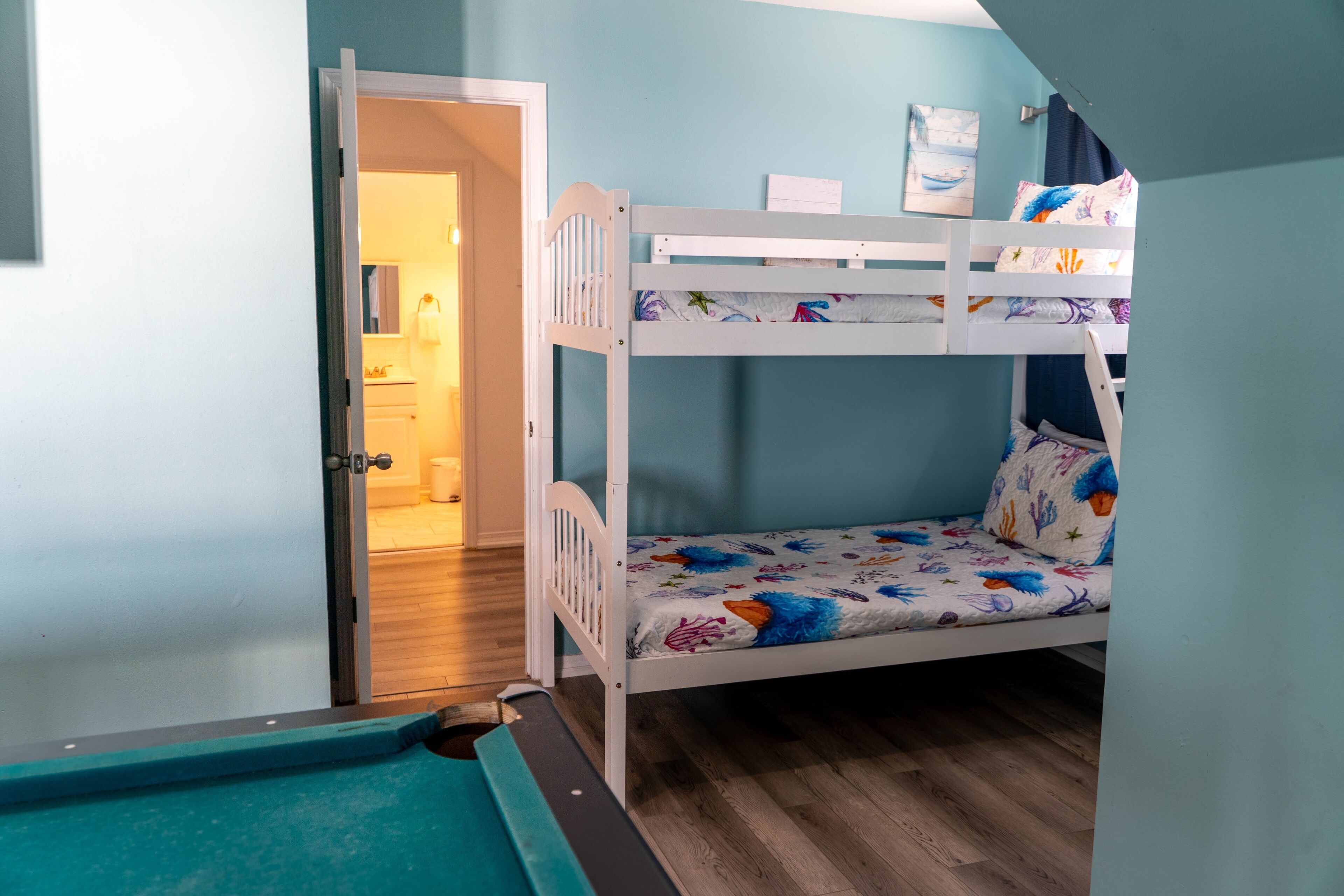 A bunk bed with a floral bedspread, a door leading to a bathroom, and a pool table in a room with teal walls.