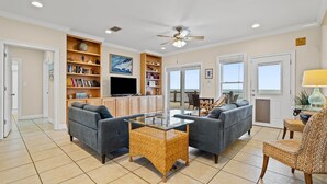 A spacious living room with a glass coffee table, two grey sofas, a bookshelf, and a television. - Off The Hook - Very Private Lot With Amazing Gulf Views Perfect For Your Family Beach Vacation 4 Bedroom Home by RedAwning (Dauphin Island)