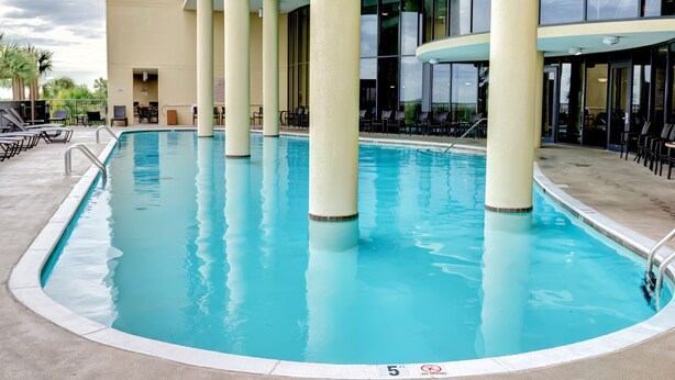 A swimming pool with clear blue water, surrounded by lounge chairs and tables, and a building with large windows in the background. - Calm Seas - Holiday Isle #119 - Tremendous Views, A Beautiful Gulf Front Pool, A Heated Indoor Pool, Jacuzzi, Steam Room, And Gulf View Fitness Center. 2 Bedroom Condo by RedAwning (Dauphin Island)