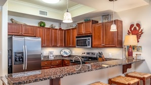 A kitchen with wooden cabinets, granite countertops, stainless steel appliances, and bar stools. - Calm Seas - Holiday Isle #119 - Tremendous Views, A Beautiful Gulf Front Pool, A Heated Indoor Pool, Jacuzzi, Steam Room, And Gulf View Fitness Center. 2 Bedroom Condo by RedAwning (Dauphin Island)