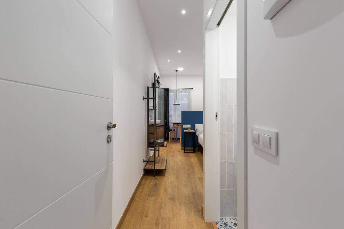 A narrow hallway with white walls and wooden flooring leading to a room with a bed and a desk.