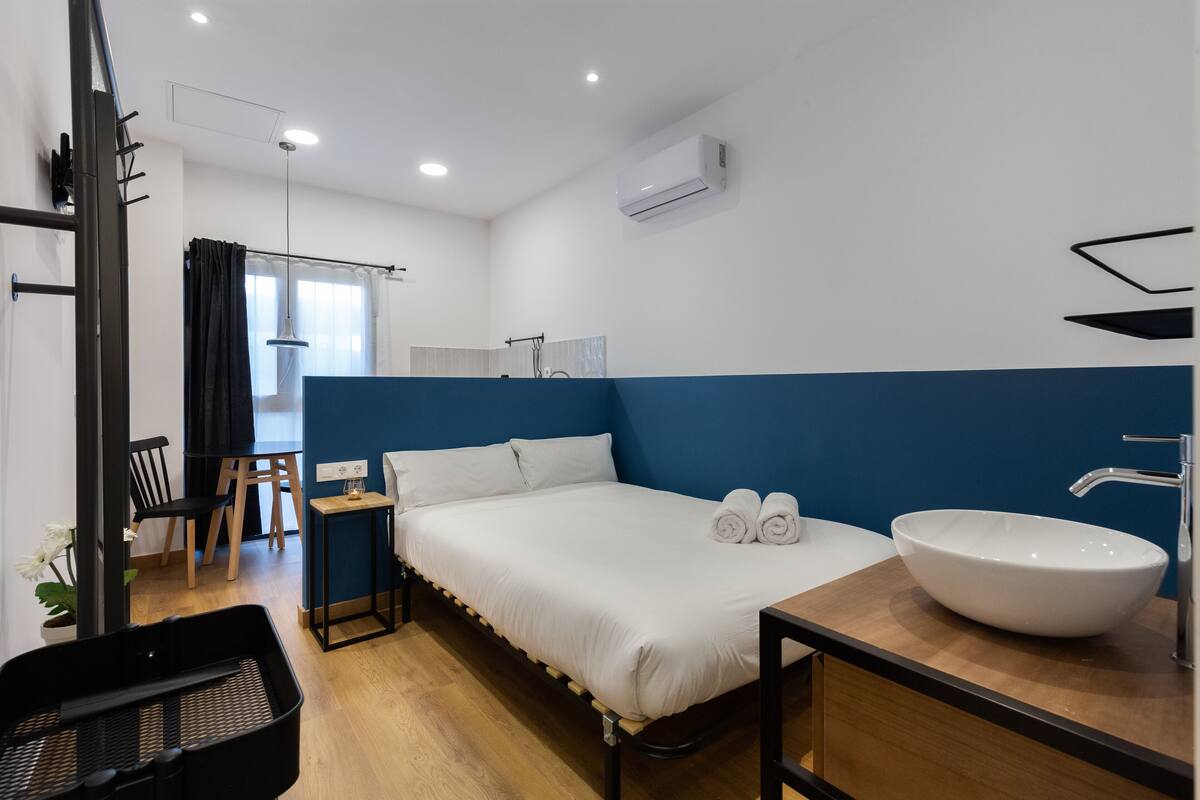 A modern hotel room with a bed, a large window, a desk, and a bathroom with a sink.