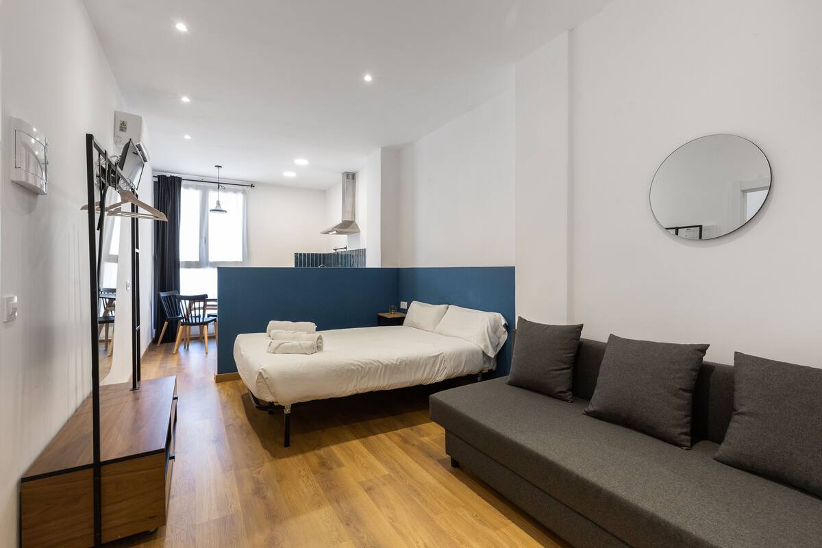 A modern hotel room with a bed, a sofa, a wooden floor, a mirror, and a window.