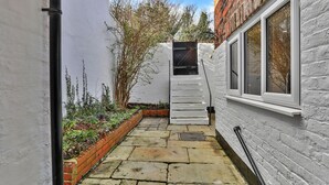 A narrow stone pathway leading to a white door with a small window, surrounded by a brick wall and vegetation.