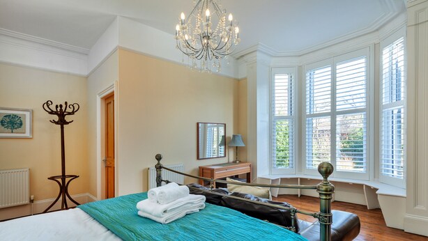 A well-lit bedroom with a large bed, a chandelier, a window with blinds, and a view of greenery outside.