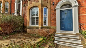 A traditional brick building with a blue door, white trim, and a small garden area with stone steps leading to the entrance.