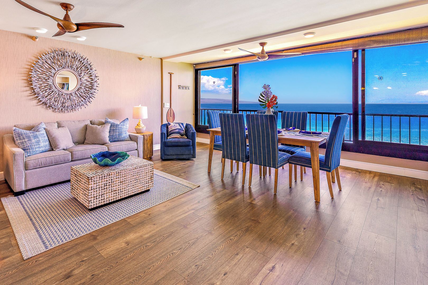 Ein geräumiges Wohnzimmer mit großem Fenster zum Meer, einer gemütlichen Sitzecke inklusive Sofa und Sesseln sowie einem Esstisch mit blau gestreiften Stühlen.