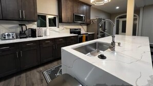 A modern kitchen with dark wooden cabinets, a white marble countertop, a stainless steel sink, and a built-in microwave. - Minutes from Anything Downtown Indy! (Indianapolis)