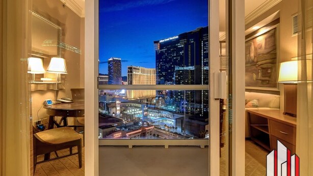 A hotel room with a large window offering a nighttime city view, a desk with a lamp, and a chair. - MGM Signature-  Studio F1 Track Strip View Balcony (Las Vegas)