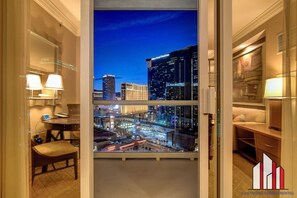 A hotel room with a large window offering a nighttime city view, a desk with a lamp, and a chair.