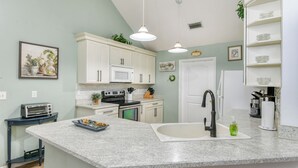 Una cocina con gabinetes blancos, encimera de granito, estufa negra, pileta blanca e iluminación colgante.