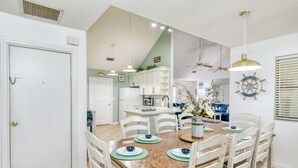 Un comedor con una mesa de madera, sillas blancas y manteles individuales azules y verdes. Se observa una cocina al fondo con armarios blancos y un sofá azul.