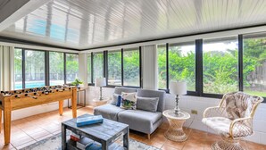 Una habitación amplia con mesa de fútbol, un sofá gris, sillas de mimbre y grandes ventanales que ofrecen vistas a la vegetación.