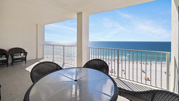 En balkon med et glasbord og fletstole, der giver udsigt over en strand og et klart hav.