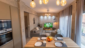 Une cuisine moderne dotée d’un four encastré, un coin repas avec une table en bois pour quatre personnes, et un salon avec un canapé et un téléviseur.