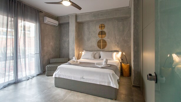 Une chambre à coucher moderne, dotée d’un grand lit, d’un ventilateur de plafond et offrant une vue sur la ville à travers de fines rideaux.