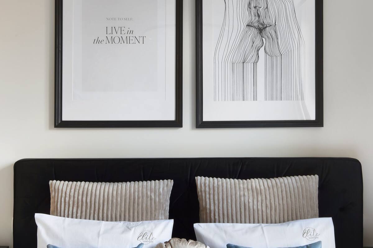A bedroom with a black headboard, two framed artworks, and a bed with white bedding and blue and beige pillows.