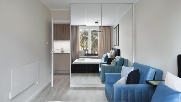 A modern hotel room with a bed, a seating area with blue sofas, a dining table, and a view of trees through the window. - Elite Apartments Garncarska (Gdansk)