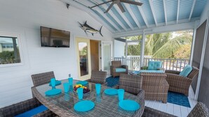 A covered outdoor dining area with wicker furniture and blue cushions, a television mounted on the wall, and a view of palm trees through large windows.