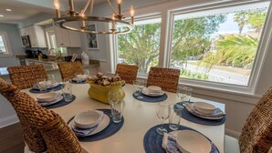A dining area with a white table set for six, surrounded by wicker chairs, a chandelier, and a view of the outdoors.