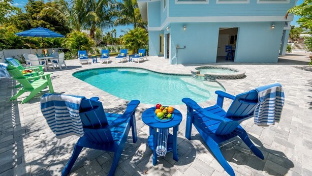 A pool area with blue and green lounge chairs, a table with fruit, and a light blue building in the background.