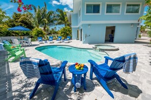 Zona de piscina con sillas reclinables azules y verdes, una mesa con fruta, y un edificio azul claro al fondo.