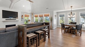 Uma sala de estar ampla com teto alto, piso de madeira, uma TV grande, uma mesa de jantar com cadeiras e um espaço bar com banquetas.