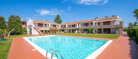A swimming pool with a brick deck, a building with multiple balconies, and a clear blue sky.