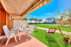 Une terrasse couverte avec une table dressée pour les repas, des chaises d’extérieur et une vue sur une piscine et une maison en arrière-plan.