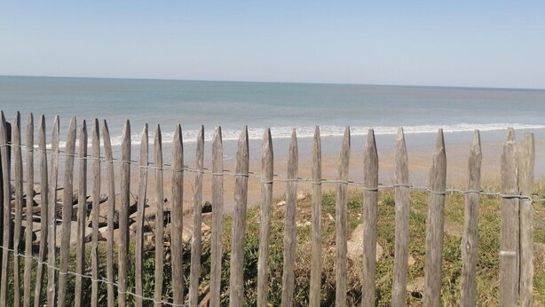 Una valla de madera con alambre de púas frente a una playa con olas tranquilas y cielo despejado.