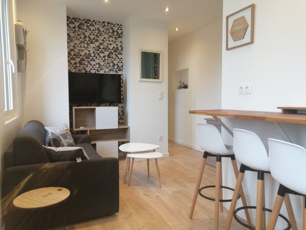 A modern living room with a sofa, a TV, a small dining table with chairs, and a patterned wall. - Le Courlis, two minutes from the beach (Hauteville-sur-Mer)