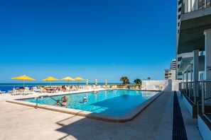A swimming pool with yellow umbrellas, a clear blue sky, and a view of the ocean.