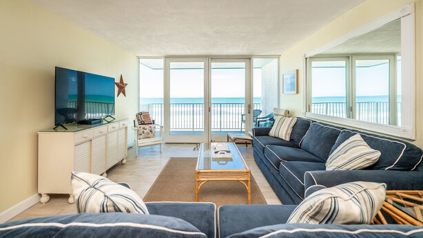A living room with a sofa, coffee table, TV, and a view of the ocean through large windows.