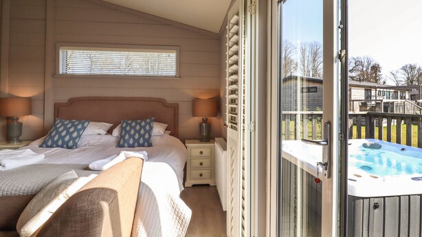 A bedroom with a large bed, two bedside lamps, a window with blinds, and a sliding glass door leading to an outdoor hot tub area.
