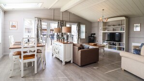A spacious living area with a high ceiling, wooden flooring, a dining table with chairs, a sofa, a TV, and a bookshelf.