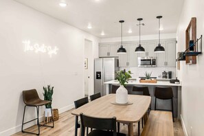 A modern kitchen with white cabinets, a stainless steel refrigerator, and a microwave. There is a dining area with a wooden table and black chairs.