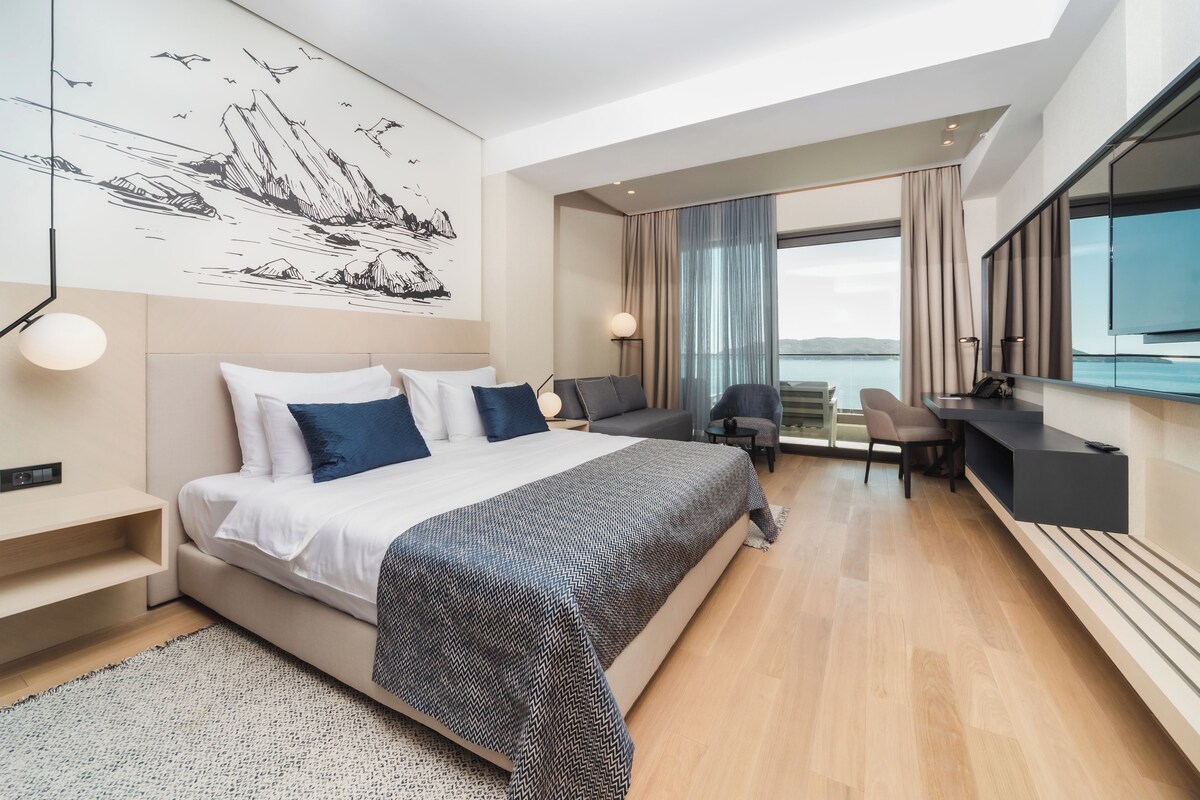 A modern hotel room with a large bed, a desk, and a view of the sea.