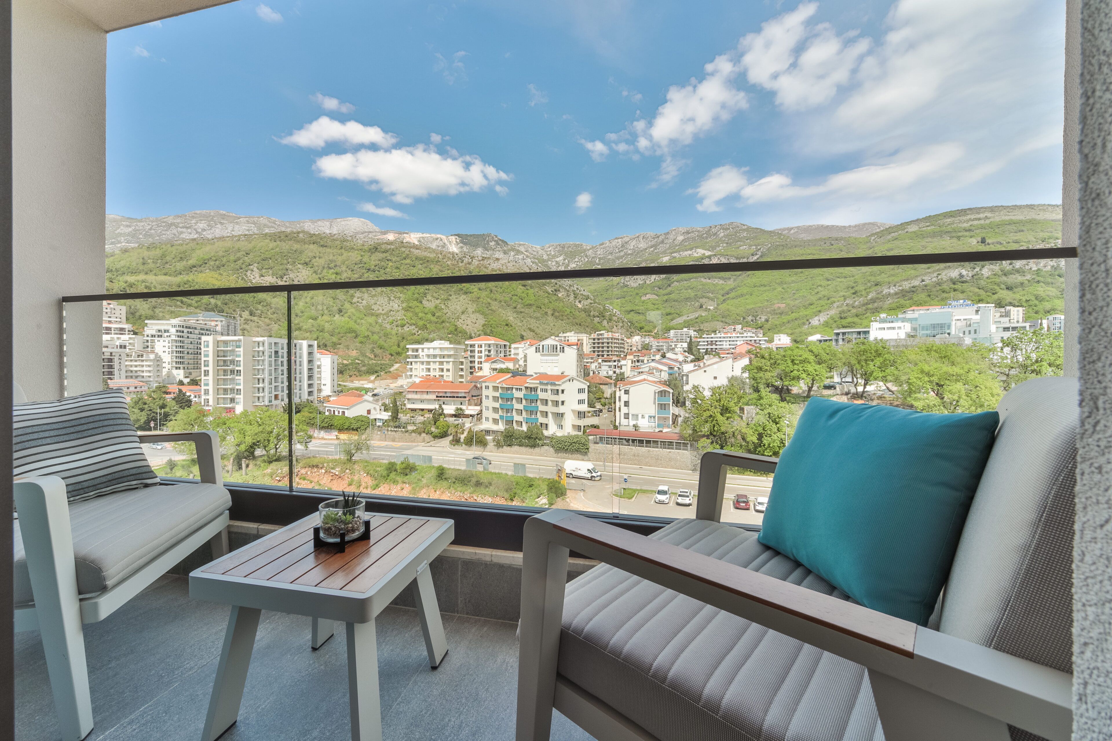 Un balcon avec un canapé et une petite table, d’où l’on peut admirer un paysage urbain composé d’immeubles et de collines.