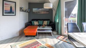 Una sala de estar moderna con un sofá negro, un otoman colorido, una mesa de centro y vistas al exterior a través de un ventanal con cortinas verdes.