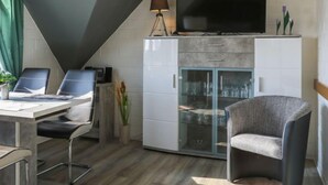 Una habitación moderna con una televisión sobre un mueble, un escritorio con sillas y una vitrina con estantes.