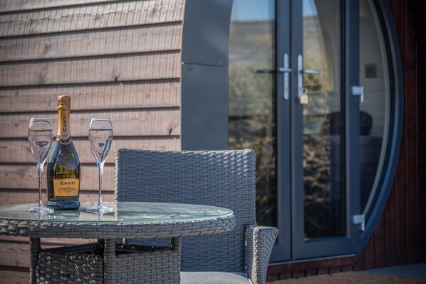 A glass table with a bottle of Cantina Prosecco and two glasses, set against a wooden wall with a large window. - Bracken Burrows - 1 Bed Pod - New Hedges (Tenby)