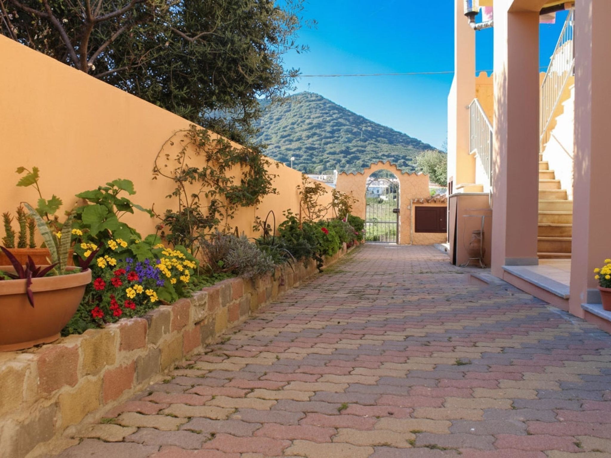 A paved pathway leading to a gate, flanked by potted plants and a hillside in the background.