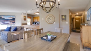 A dining area with a wooden table, chairs, a chandelier, a clock, and a kitchen in the background. - Ski-in condo located 1 block from Main St w/ hot tubs, Covered parking, Fantasti (Breckenridge)