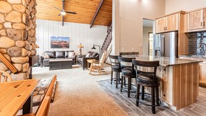 A spacious living area with a stone wall, wooden ceiling, and a kitchen with wooden cabinets and a large island. - Fireplace, Private Balcony, River/Forest Views! (Breckenridge)