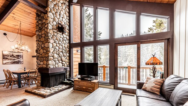 A cozy living room with a stone fireplace, a television, and a large window overlooking a deck. - Fireplace, Private Balcony, River/Forest Views! (Breckenridge)