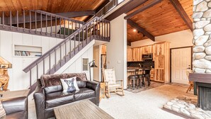 A spacious living area with a leather sofa, a wooden coffee table, and a stone fireplace. A wooden staircase leads to an upper level. - Fireplace, Private Balcony, River/Forest Views! (Breckenridge)
