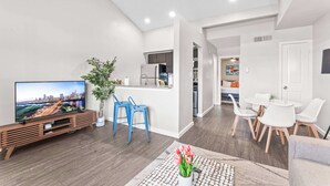 Una sala moderna con televisor de pantalla plana, mueble de madera para entretenimiento, sillas azules, una zona de comedor con sillas blancas y una cocina al fondo.
