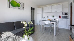 A modern living room with a sofa, a dining table, and chairs. There is a kitchen area with white cabinets and appliances.
