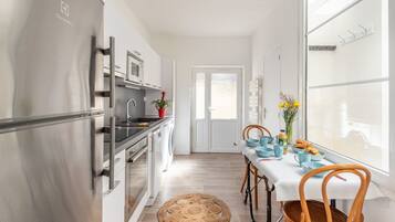 Une cuisine moderne équipée d’appareils électroménagers en acier inoxydable, un coin repas avec une table dressée pour deux personnes et une vue dégagée sur l’extérieur grâce à une grande fenêtre.
