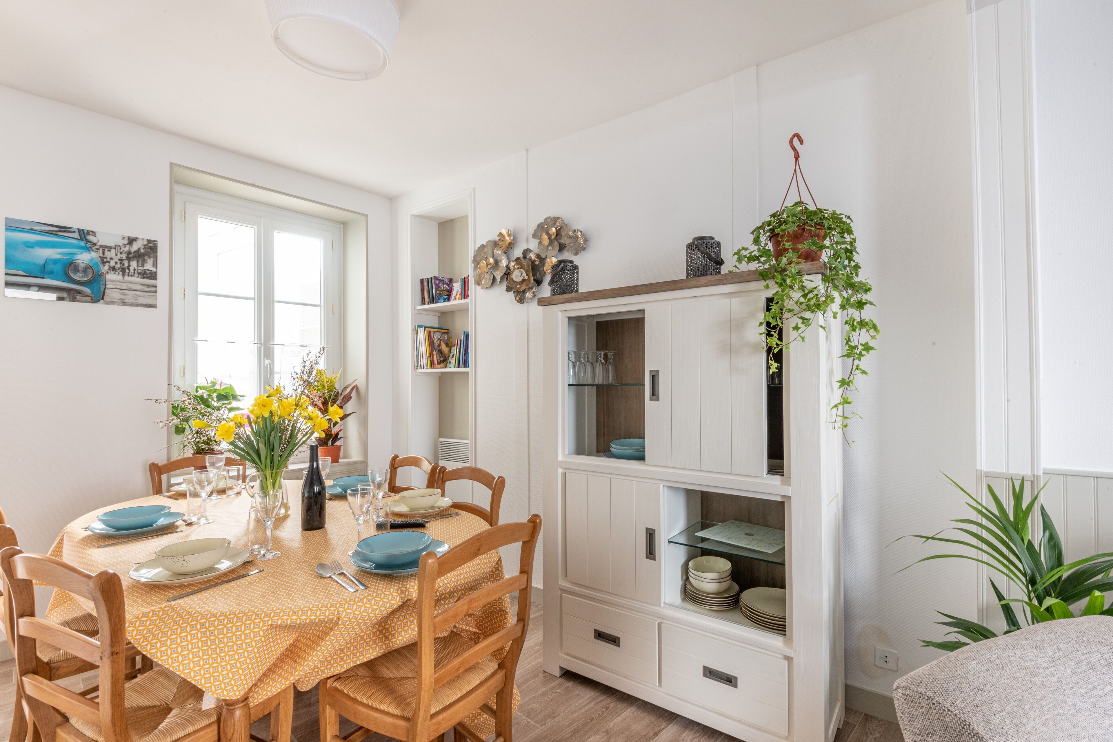 Een eethoek met een ronde tafel gedekt voor vier personen, omringd door houten stoelen. Aan de rechterkant staat een witte kast met glazen deuren, en erboven hangt een kamerplant.
